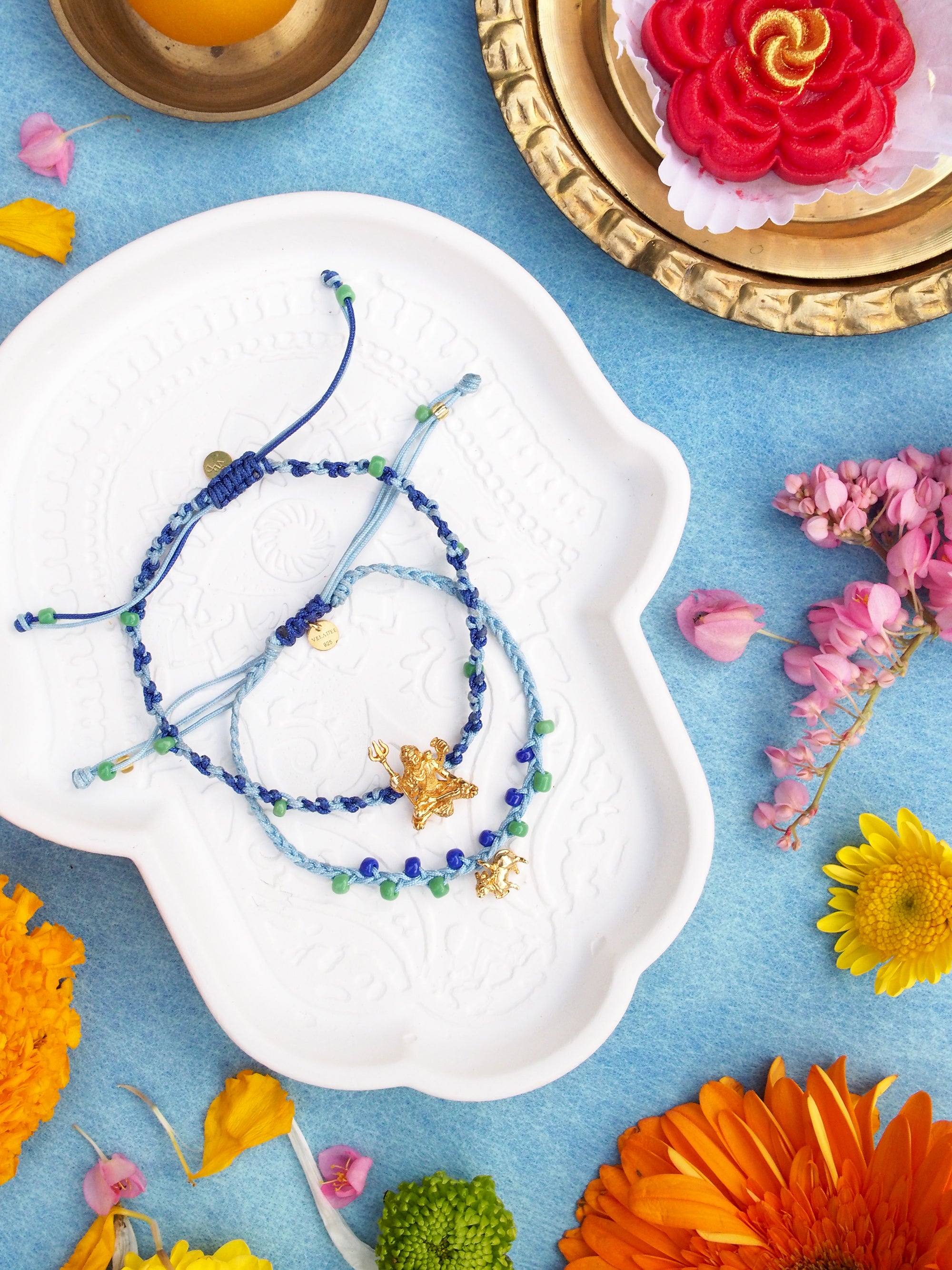Two colorful bracelets, featuring a gold Trimurti Shiva charm and a gold Nandi charm surrounded by festive sweets and vibrant flowers.