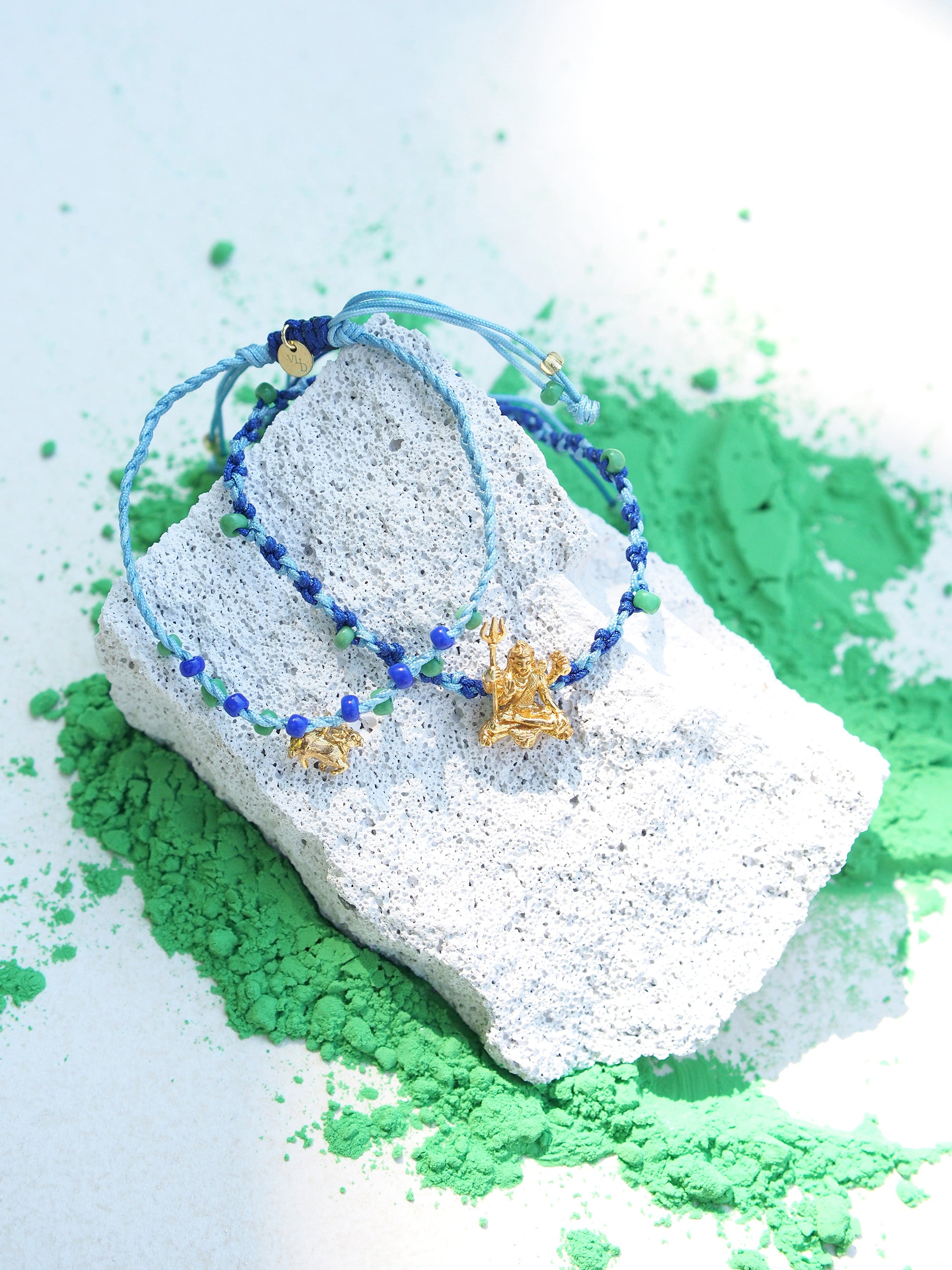Two colorful bracelets in blue threads with blue and green beads, featuring a gold Trimurti Shiva charm and a gold Nandi charm placed on a textured surface among green powder.