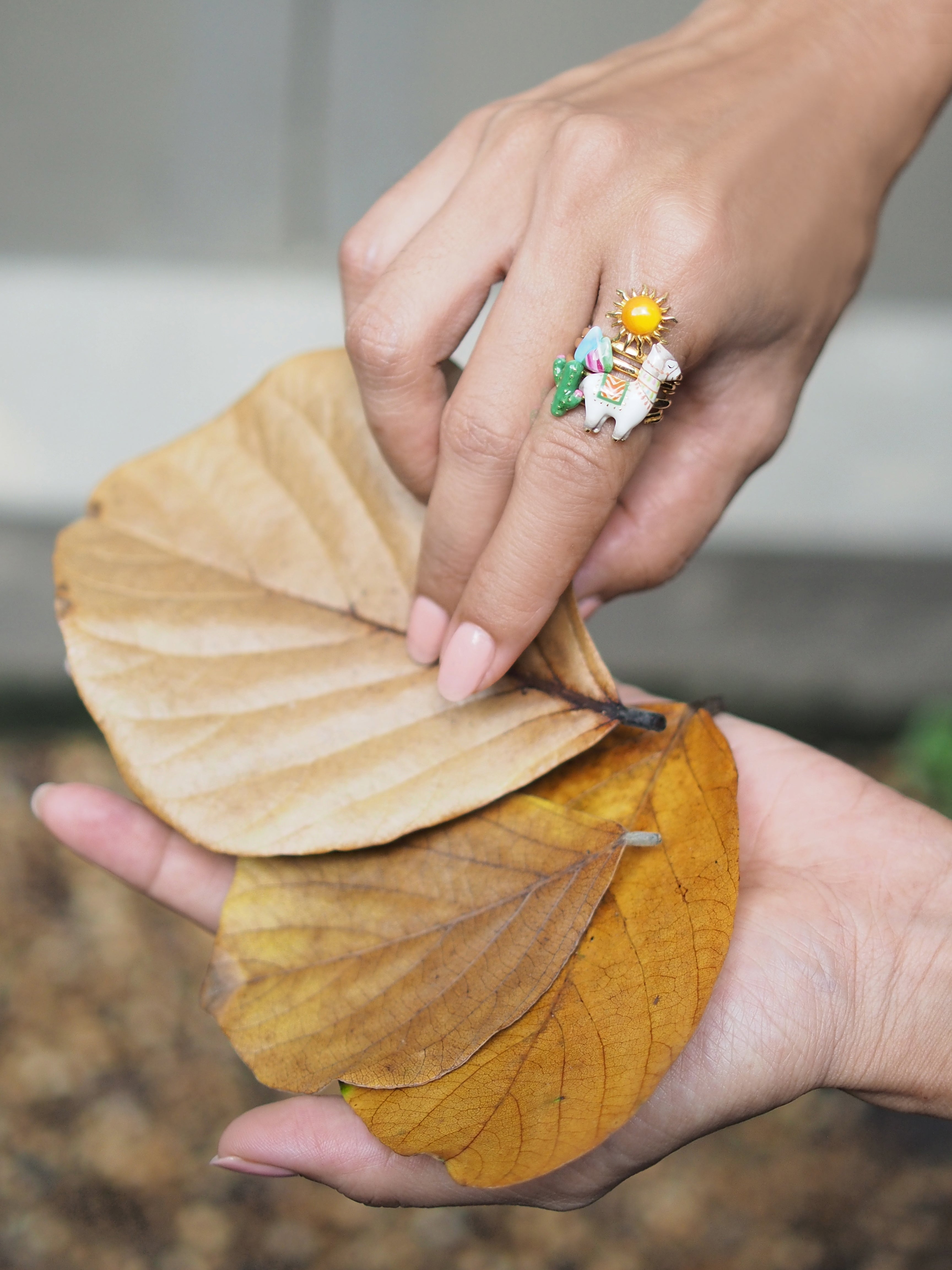 Artistic stacking ring featuring a sun, alpaca, cactus, and mountain design, worn on a hand holding autumn leaves.