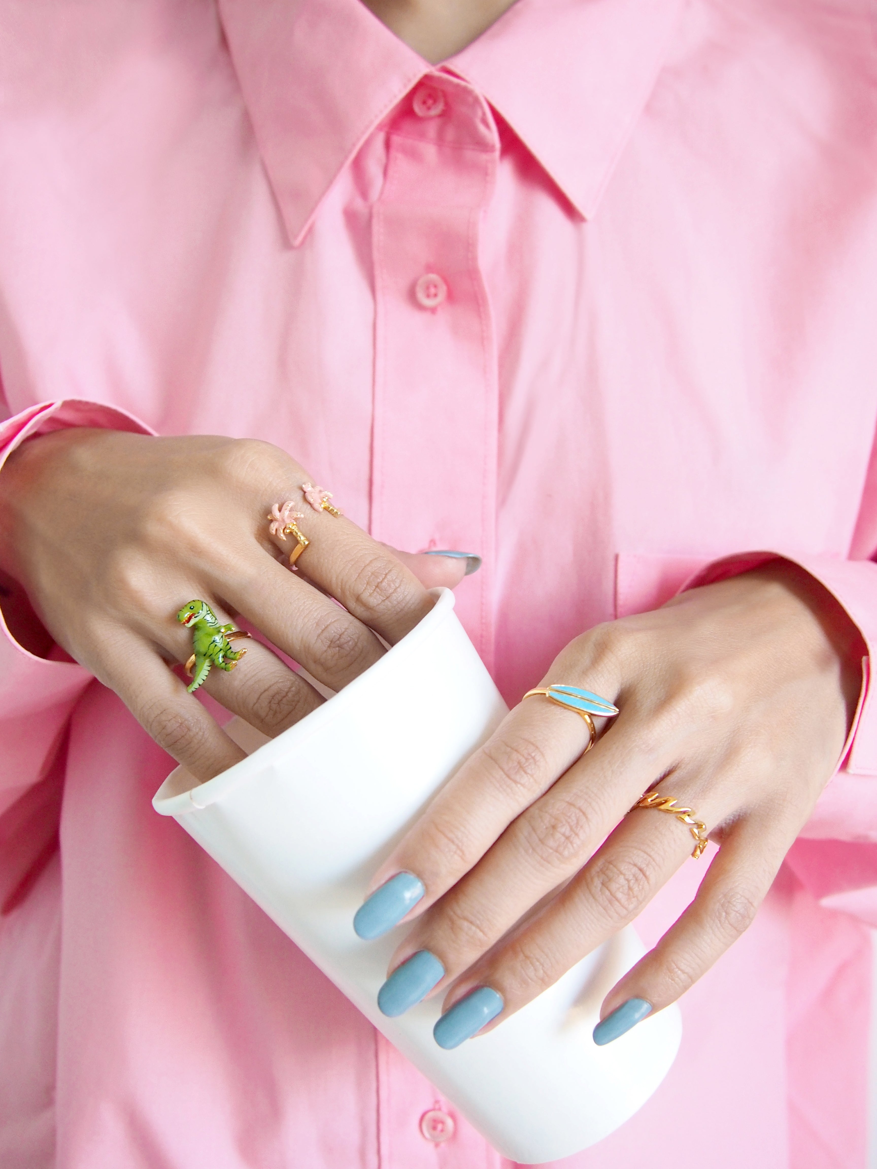 Two hand adorned with cute rings featuring a T-rex, palm trees, a surfboard, and a wave-pattern design holding a white cup in a pink shirt.